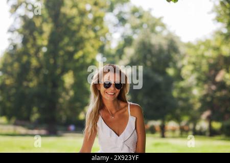 Una giovane donna allegra che indossa occhiali da sole e un vestito bianco sorride mentre si trova in un parco verde lussureggiante in una giornata di sole. Foto Stock