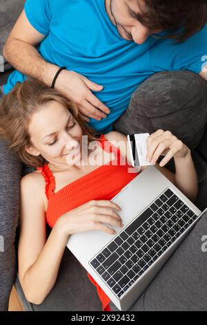 Un giovane uomo e una donna, comodamente sdraiati su un divano grigio, utilizzano un computer portatile per fare acquisti online, tenendo insieme una carta di credito. Foto Stock