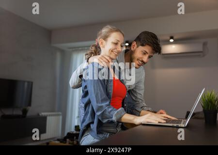 Una giovane coppia allegra che si occupa di un notebook nel loro accogliente salotto, condividendo un momento di relax insieme. Foto Stock