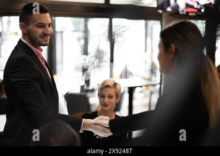 Un gruppo diversificato di giovani professionisti stringe la mano in una caffetteria illuminata da calore, suggerendo lavoro di squadra ed espansione della rete. Foto Stock