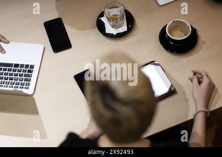 Vista dall'alto di una riunione d'affari in un bar moderno, con le mani che usano computer portatili e smartphone, accompagnato da un caffè. Foto Stock