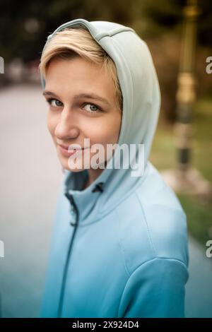 Un ritratto ravvicinato di una giovane donna caucasica che indossa una felpa con cappuccio blu, sorridendo dolcemente in un parco all'aperto. Foto Stock