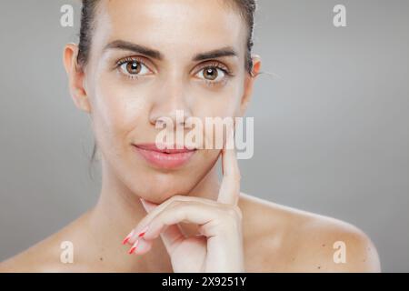 Questa immagine cattura una giovane donna con pelle limpida e trucco naturale, toccando delicatamente la guancia, trasudando fiducia ed eleganza. Foto Stock