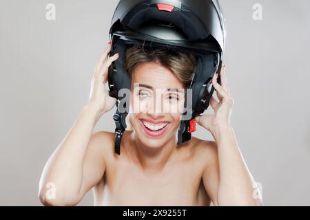 Una giovane e allegra donna sorride mentre regola il suo casco da moto nero, incarnando un senso di sicurezza, divertimento e avventura in un ambiente da studio. Foto Stock