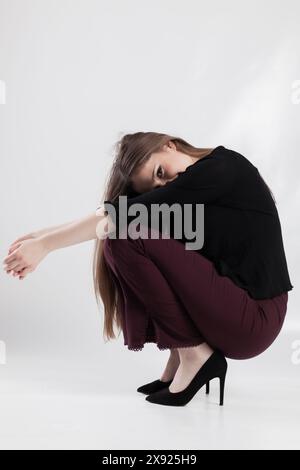 Una giovane donna elegante si trova in una posa suggestiva, indossando una camicetta nera, pantaloni color marrone e tacchi alti su uno sfondo bianco. Foto Stock