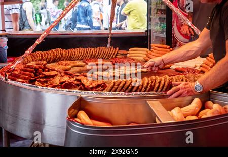 ampia griglia ripiena di vari tipi di salsicce, polpettine e carne cucinata. Una persona tende a grigliare, utilizzando pinze per preparare la salsiccia. La S Foto Stock