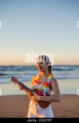 la giovane donna multiculturale verticale ama giocare l'ukulele sulla costa durante un bellissimo tramonto estivo, creando un'atmosfera tranquilla con la s. Foto Stock