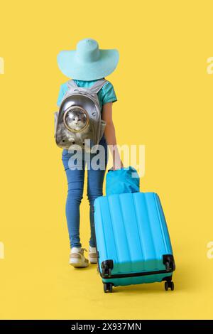 Viaggiatrice donna con gatto nel portabagagli e valigia su sfondo giallo, vista posteriore Foto Stock