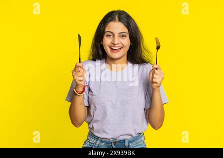 Pronti a mangiare. Una donna indiana affamata che aspetta di servire piatti per la cena con un coltello da forchetta per posate senza irrequietezza apprezzerà il delizioso pasto del ristorante. Ragazza eccitata su sfondo giallo Foto Stock