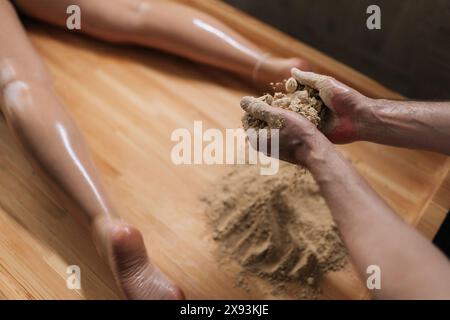 Vista dall'alto dell'irriconoscibile massaggiatore ayurvedico che mescola sabbia riscaldata sul lettino per massaggi per trattamenti di scrubbing in un lussuoso salone spa. Foto Stock