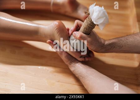 Vista dall'alto di una giovane donna irriconoscibile e rilassata che riceve un massaggio ai piedi ayurvedico con borse alle erbe Kizhi di un massaggiatore maschile nel salone spa del resort di lusso. Foto Stock