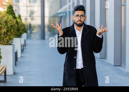 Cosa? Perché? Sorpreso un uomo d'affari indiano frustrato che alza la mano chiedendo il motivo del fallimento, dimostrando incredulità irritazione da problemi all'aperto. Confuso ragazzo indù arabo libero professionista in città Foto Stock