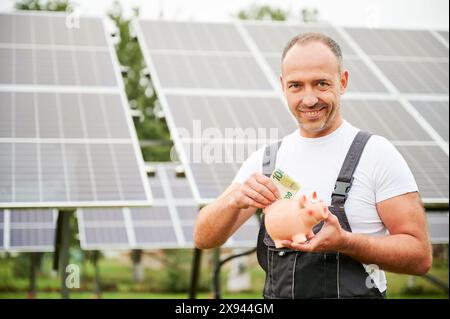 Uomo esperto che investe nell'energia verde per profitti futuri. Adulto maschio felice dell'investimento che ha fatto. Uomo in tuta che mette euro al salvadanaio sullo sfondo di una batteria solare. Foto Stock