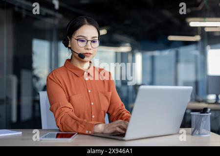 Operatore di contact centre che indossa un auricolare mentre lavora diligentemente su un computer portatile presso una moderna scrivania. Ambiente di lavoro professionale. Foto Stock