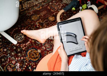 Parigi, Francia - 11 giugno 2023: Una donna è seduta e sta navigando sul sito Web di Apple su un iPad, visualizzando i dettagli delle cuffie Vision Pro, con i piedi appoggiati su una sedia e i giocattoli per bambini sparsi sul pavimento Foto Stock