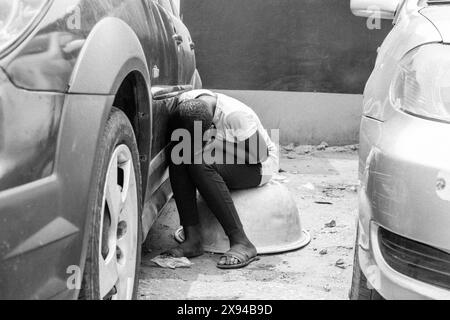 Una giovane donna che riposa tra le auto parcheggiate in Ghana, seduta su una grande ciotola, cattura un momento di stanchezza tra le attività quotidiane. Foto Stock