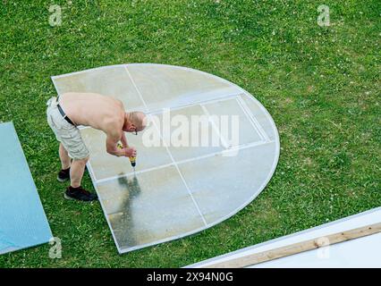 L'uomo assembla il kit per la serra domestica a casa. I pezzi sono disposti intorno all'erba del giardino di casa e si adattano insieme, sopra la vista. Foto Stock