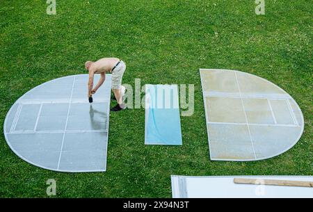 L'uomo assembla il kit per la serra domestica a casa. I pezzi sono disposti intorno all'erba del giardino di casa e si adattano insieme, sopra la vista. Foto Stock