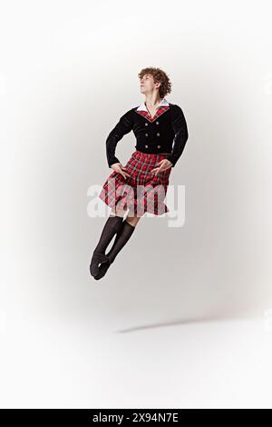 Il giovane, ballerino, vestito con kilt rosso tartan e giacca nera, posa con fermezza e precisione su sfondo bianco dello studio. Foto Stock