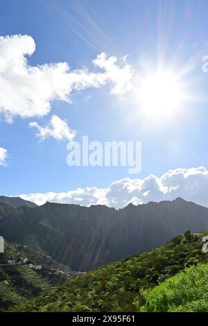 Il sole splende in modo luminoso sulle verdi colline e sulle imponenti cime delle montagne, con nuvole sparse in un cielo blu vivace Foto Stock