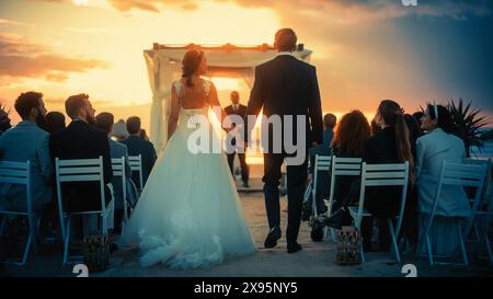 Splendida sposa in abito da sposa bianco e bel guardaroba in abito nero che scende lungo la navata in una cerimonia all'aperto vicino al mare al tramonto. Felici amici multietnici che celebrano il matrimonio. Foto Stock