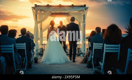 Splendida sposa in abito da sposa bianco e bel guardaroba in abito nero che scende lungo la navata in una cerimonia all'aperto vicino al mare al tramonto. Felici amici multietnici che celebrano il matrimonio. Foto Stock