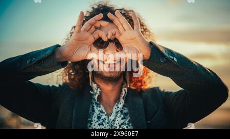 Ritratto di un giovane adulto felice con i capelli ricci lunghi, con occhiali da sole a forma di cuore, orecchini e anello del naso in posa per la fotocamera. Bell'uomo caucasico multietnico sorridente. Modifica colore caldo. Foto Stock