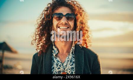 Ritratto di un giovane adulto felice con i capelli ricci lunghi, con occhiali da sole a forma di cuore, orecchini e anello del naso in posa per la fotocamera. Bell'uomo caucasico multietnico sorridente. Modifica colore caldo. Foto Stock