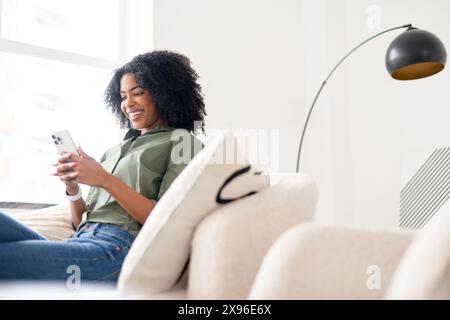 Una giovane donna afro-americana, con un sorriso affascinante, si impegna comodamente con il suo smartphone in una stanza ben illuminata, suggerendo un momento di piacevole interazione online Foto Stock