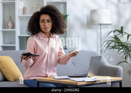 Donna stressata che gestisce le finanze personali a casa con documenti, calcolatrice e laptop, mentre è seduta su un divano in salotto. Foto Stock