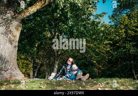 Una donna seduta sull'erba sotto un albero che tiene un bambino circondato da una foresta in autunno Foto Stock