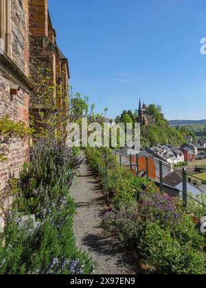 Sentiero del giardino lungo un muro di pietra con vista su un castello lontano e case su una collina sotto un cielo blu, vecchie rovine del castello sopra un fiume con alberi verdi Foto Stock