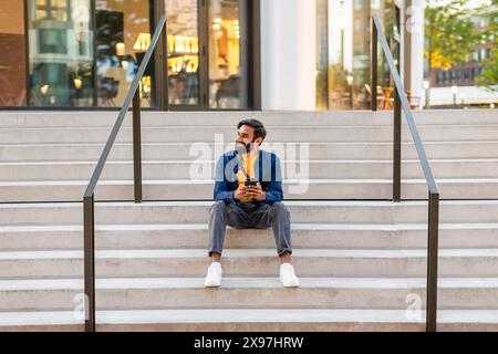 Giovane uomo seduto sulla scalinata urbana al mattino con smartphone Foto Stock