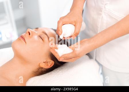 Estetista che si prepara ad applicare la schiuma detergente sulla procedura di bellezza del viso femminile, idratando la pelle del viso, massaggio facciale. Il cosmetologo pulisce la pelle con la schiuma. Foto Stock