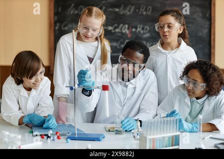 Giovane insegnante afroamericano seduto a tavola in classe dimostrando una reazione chimica al gruppo di studenti delle scuole medie Foto Stock