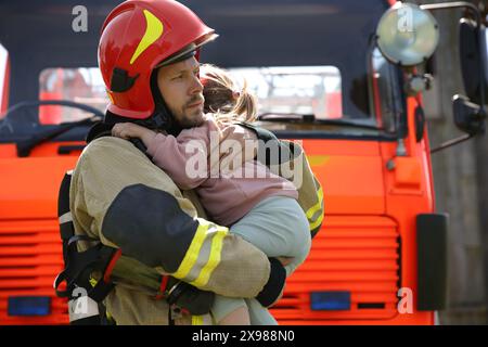 Vigile del fuoco in uniforme che tiene in mano la bambina salvata vicino al camion dei pompieri all'aperto. Salvare la vita Foto Stock