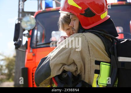 Vigile del fuoco in uniforme che tiene in mano la bambina salvata vicino al camion dei pompieri all'aperto. Salvare la vita Foto Stock