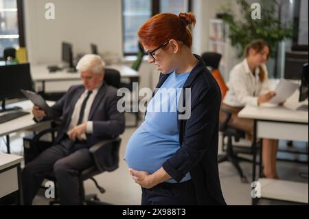 Una donna incinta soffre di dolore e tiene lo stomaco in piedi nel mezzo dell'ufficio accanto ai suoi colleghi. Foto Stock