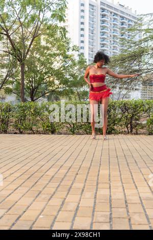 Una donna con un vivace costume da ballo rosso poggia graziosamente su un sentiero lastricato con vegetazione lussureggiante e alti edifici sullo sfondo, che mettono in mostra la città e. Foto Stock