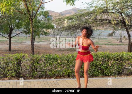 Allegra donna che balla in costume rosso all'aperto, capelli ricci, movimento espressivo. Foto Stock