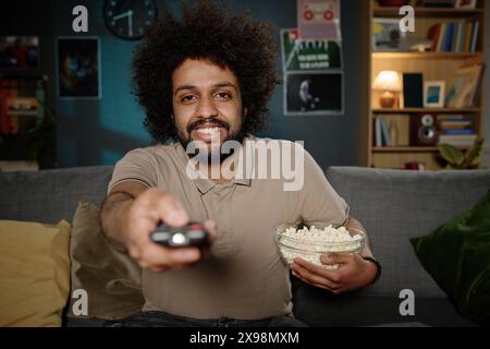 Punto di vista di un giovane con telecomando e ciotola di popcorn seduti sul divano del soggiorno per accendere la TV Foto Stock