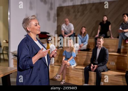 Docente femminile di abbigliamento formale sorridente, tenendo conferenze a gruppi di persone sullo sfondo, le persone si sono riunite per ascoltarla parlare di me Foto Stock