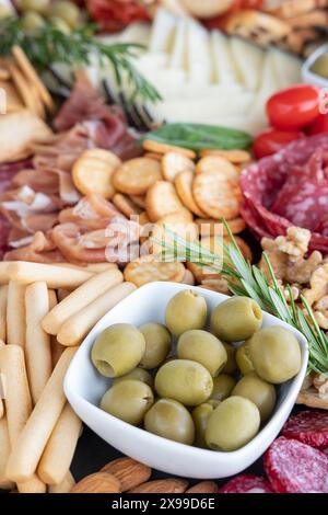 Tagliere di salumi con una ciotola in ceramica ripiena di olive verdi incastonata al centro di un'ampia selezione di prelibatezze: Affettati e fette di formaggio Foto Stock
