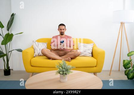 Un giovane sorridente con lo smartphone seduto sul divano di casa Foto Stock