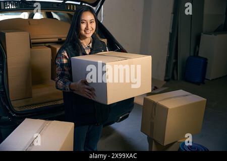 Donna felice seduta nel bagagliaio di un'auto con una scatola in mano. Foto ritagliata Foto Stock