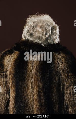 Donna anziana con capelli bianchi ricci in piedi con la schiena alla macchina fotografica, con indosso un cappotto di pelliccia, su un semplice sfondo scuro. Foto Stock