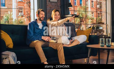 Ritratto di una giovane coppia che trascorre del tempo a casa, seduto su un divano, uomo che abbraccia la sua ragazza mentre guarda un eccitante programma televisivo in Apartment. Uomo e donna in streaming e lasciati sorprendere. Foto Stock