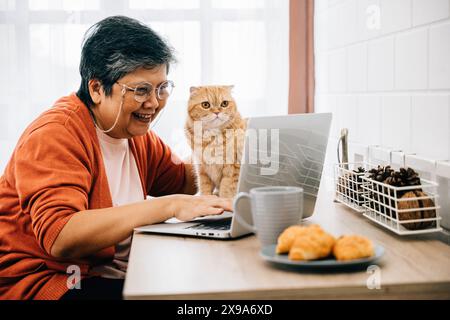 Una donna, nel suo ufficio a casa, digita il suo laptop con il suo anziano gatto appollaiato sulla scrivania. Loro Foto Stock