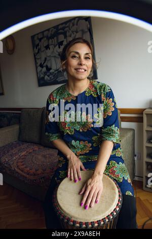 Immagine verticale, vista attraverso un anello di luce di una donna latina che suona il tamburo djembe facendo un video per le reti. Indossa un colore africano Foto Stock