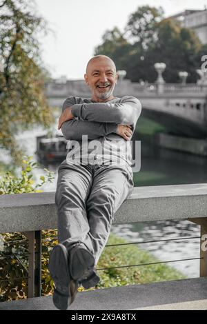 Gioioso uomo anziano in maglione grigio che si rilassa su una panchina vicino al fiume, sorridendo Foto Stock